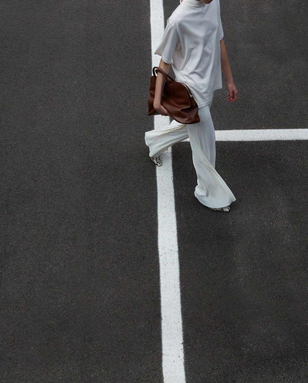 A model in a loose white outfit carries a soft brown bag while walking over white road markings